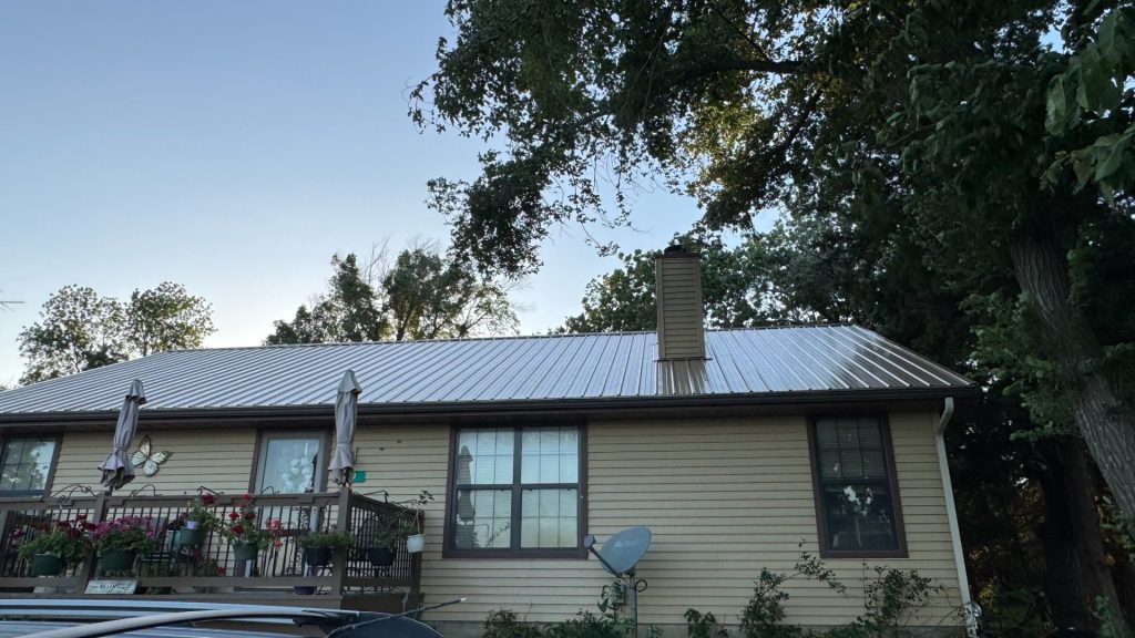 Single-family home with light beige siding, dark brown trim, a light corrugated metal roof, brick chimney, wooden deck with patio umbrellas and potted plants, and a satellite dish in a wooded setting.
