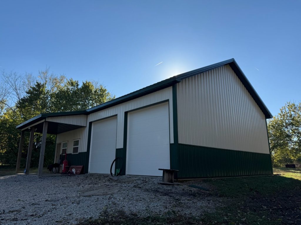 Two-tone metal pole barn or workshop with evergreen and beige vertical panels, a covered porch, two white overhead garage doors, and a walk-in entry door for durable low-maintenance use.